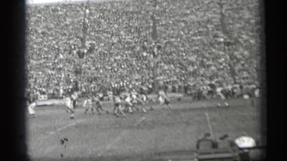 Black-and-white 8mm frame of a 1944 San Francisco football game, with players converging on the field and a large crowd in the stands.