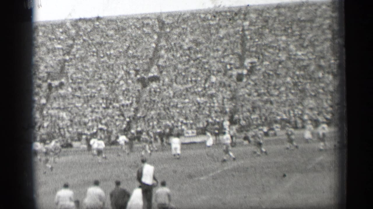 Football game viewed from the stands with players on the field and a packed stadium behind them.