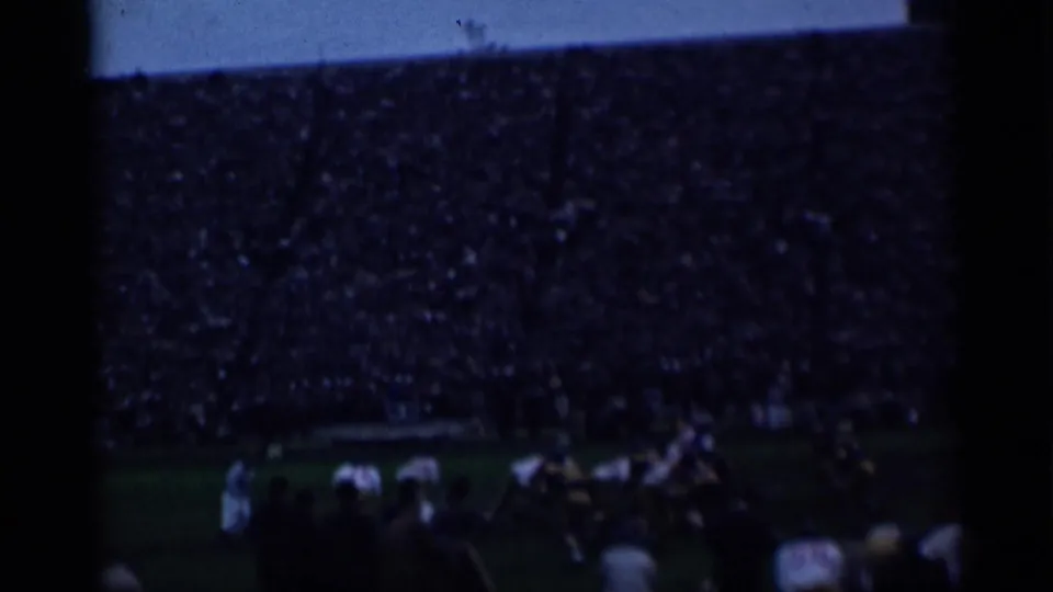 Underexposed 8mm frame from the 1944 San Francisco football footage, with players and a packed stadium visible through heavy grain.