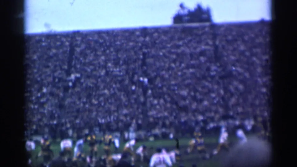 Blue-toned 8mm frame of the 1944 San Francisco football game, showing players near the sideline and a packed stadium crowd.