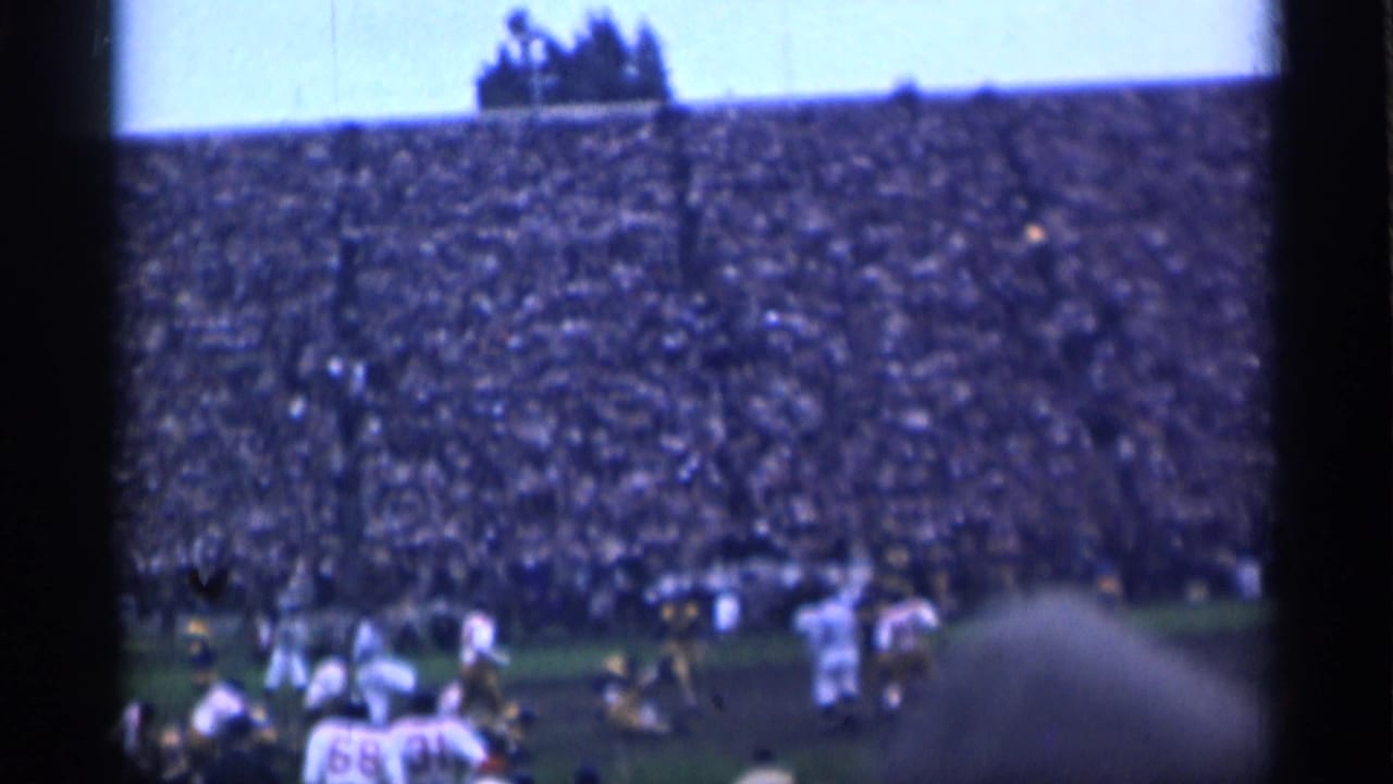 Football players on a field before a densely filled stadium grandstand.