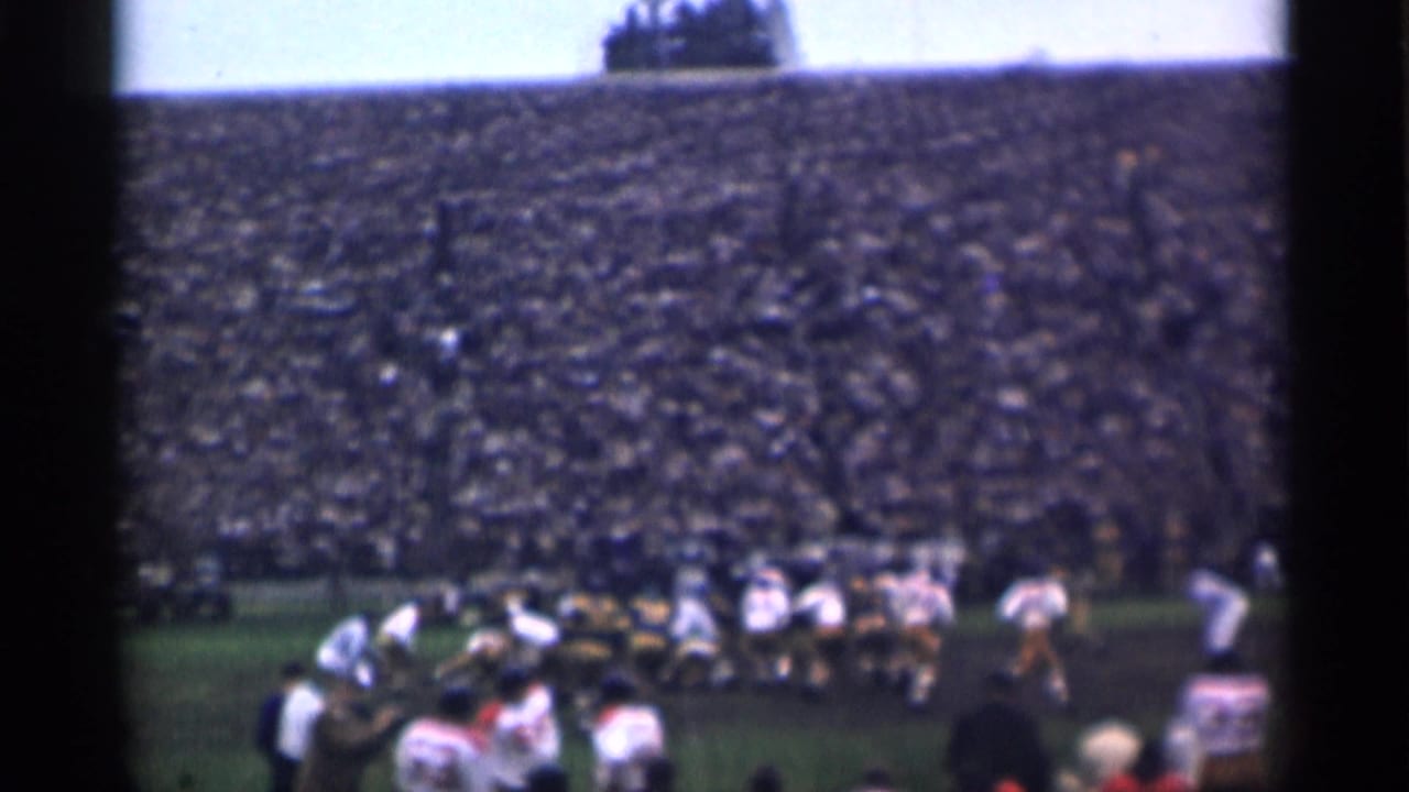 Football players line up on a field before a packed stadium crowd.
