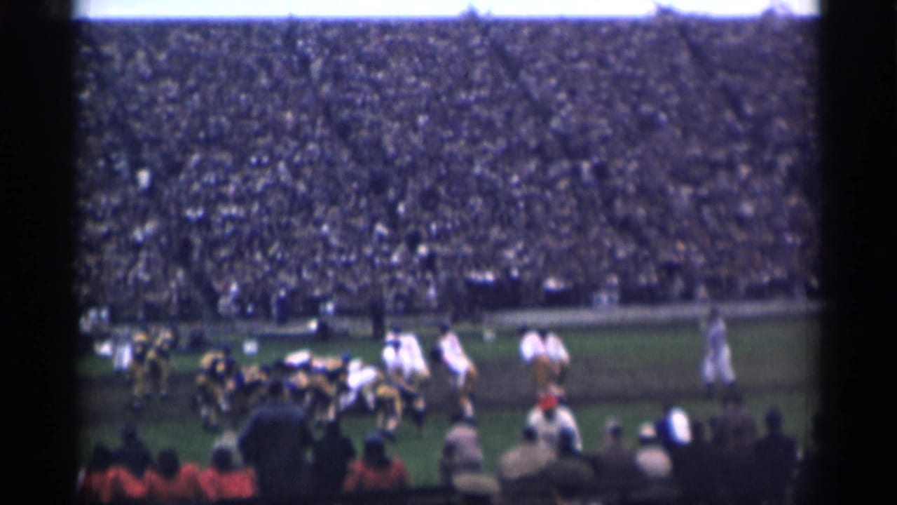 Football players line up on a field before a packed stadium crowd.