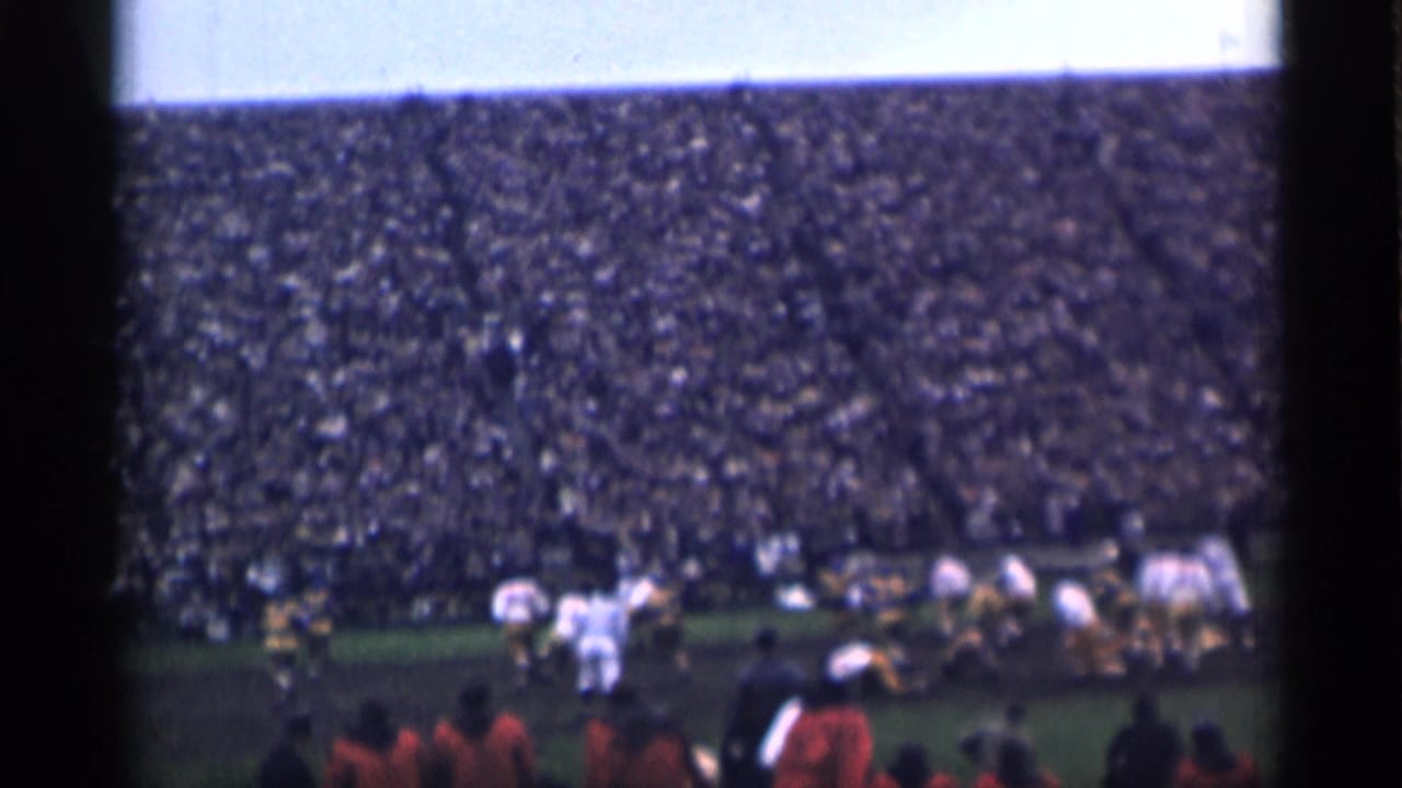 Football players on a field before packed stadium stands.