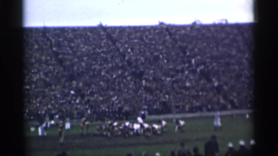 Wide 8mm frame of a football formation near the sideline, with packed stands rising behind the field.