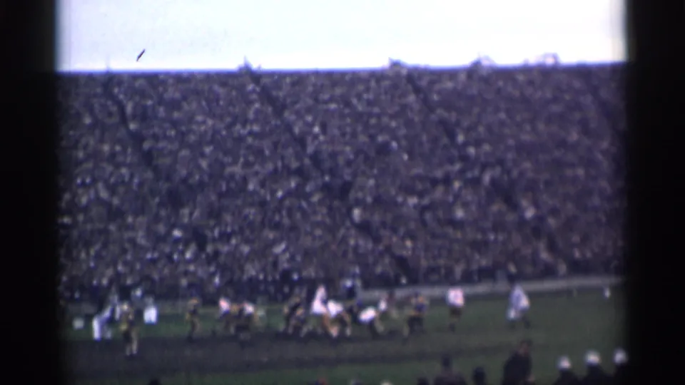 Distant home-movie view of players clustered during a football play, framed by dense stadium seating and film borders.