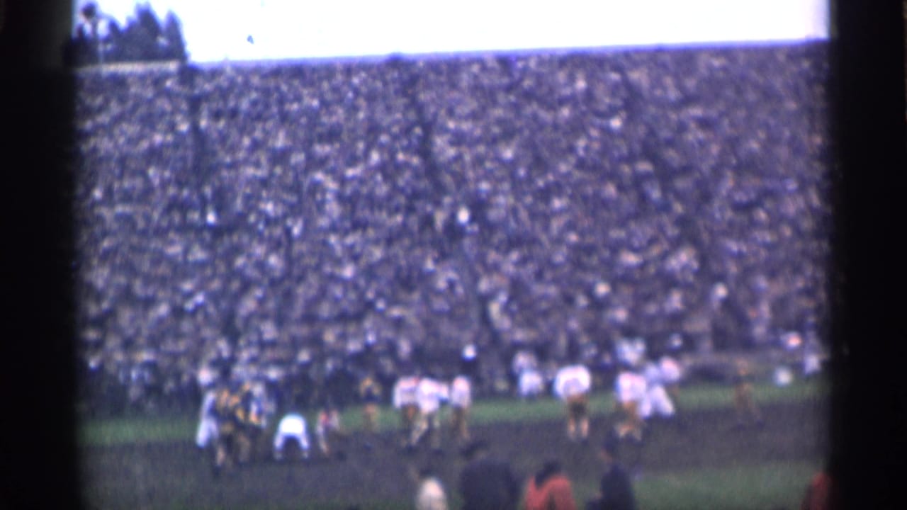 Football players line up on a field before a packed stadium crowd.