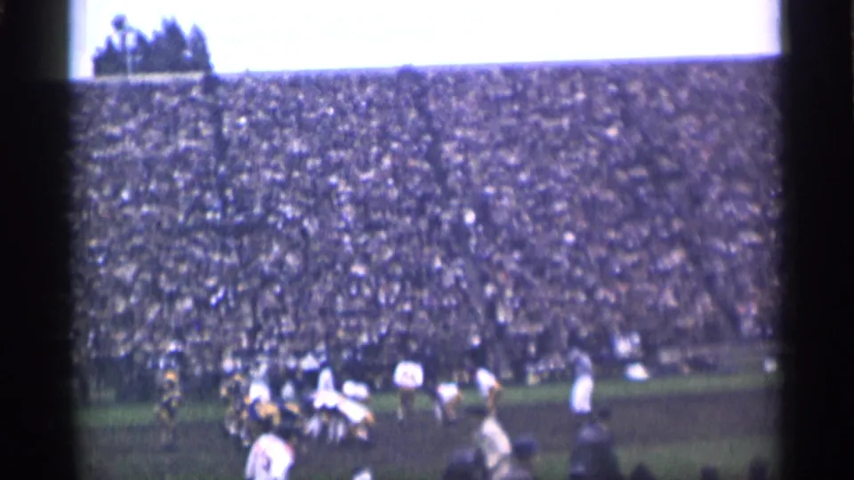 Distant 1944 San Francisco football action seen from the stands, with players converging near the center of the field and a large crowd behind them.