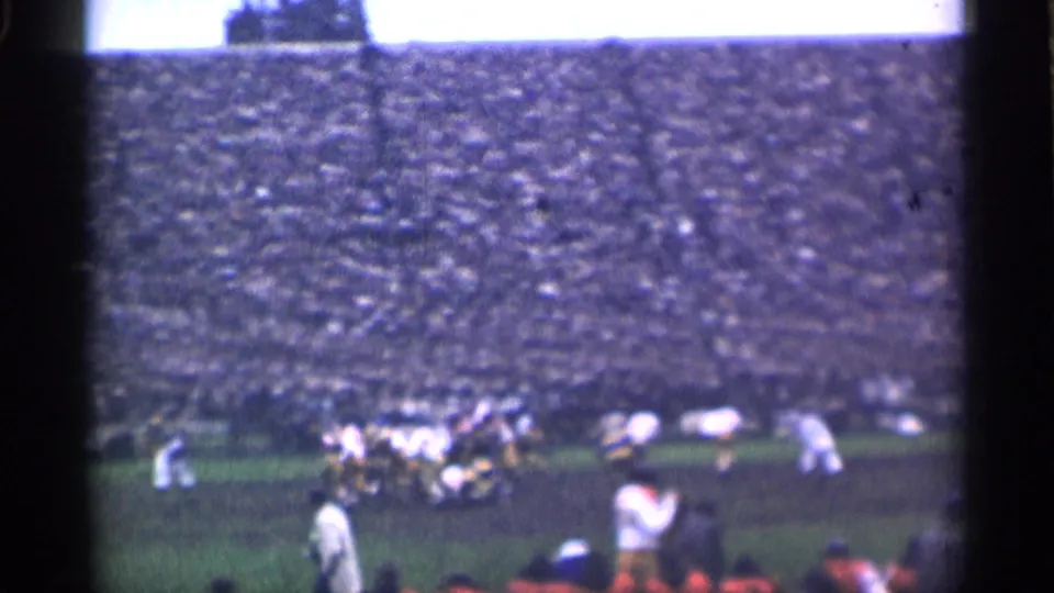Home-movie frame from a 1944 San Francisco football game, showing players near the line and foreground spectators watching from the stands.
