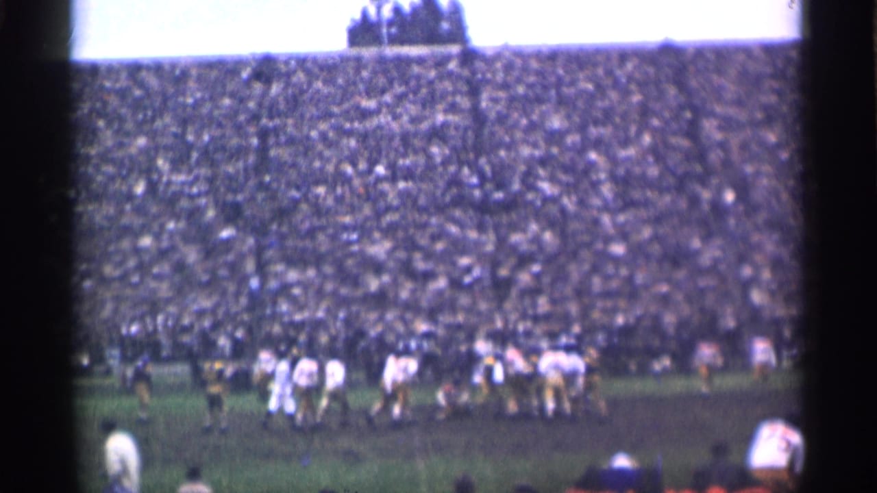 Football players line up on a muddy field before a packed stadium crowd.