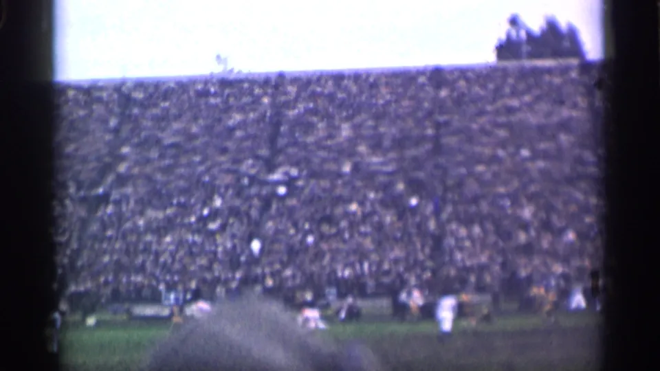 The stadium crowd dominates the frame while football action continues near the lower edge, partly blocked by nearby spectators.