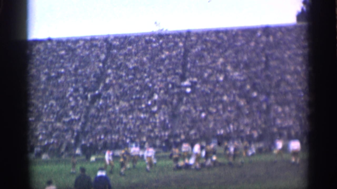 Football players on a grass field before a packed stadium crowd.