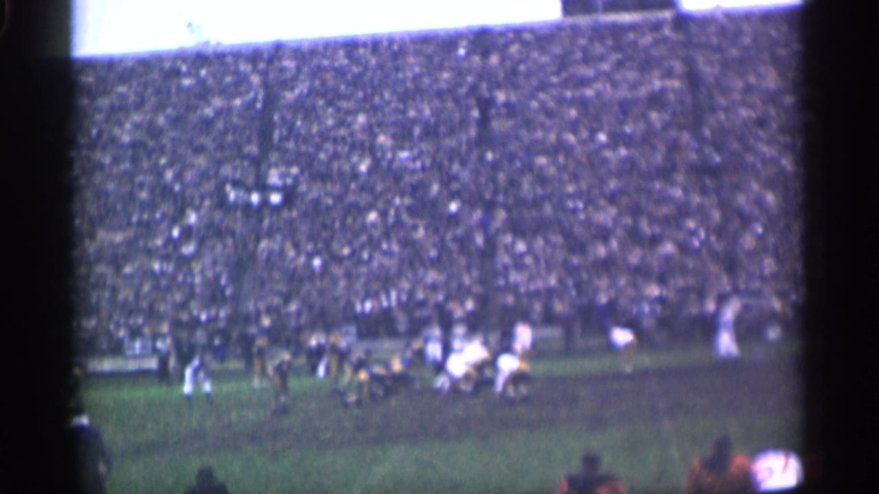 Wide view of players on a field with a packed stadium crowd behind them.