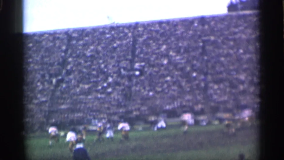 Wide 8mm frame of the San Francisco rugby match, with players in motion across the lower field and crowded stands above.