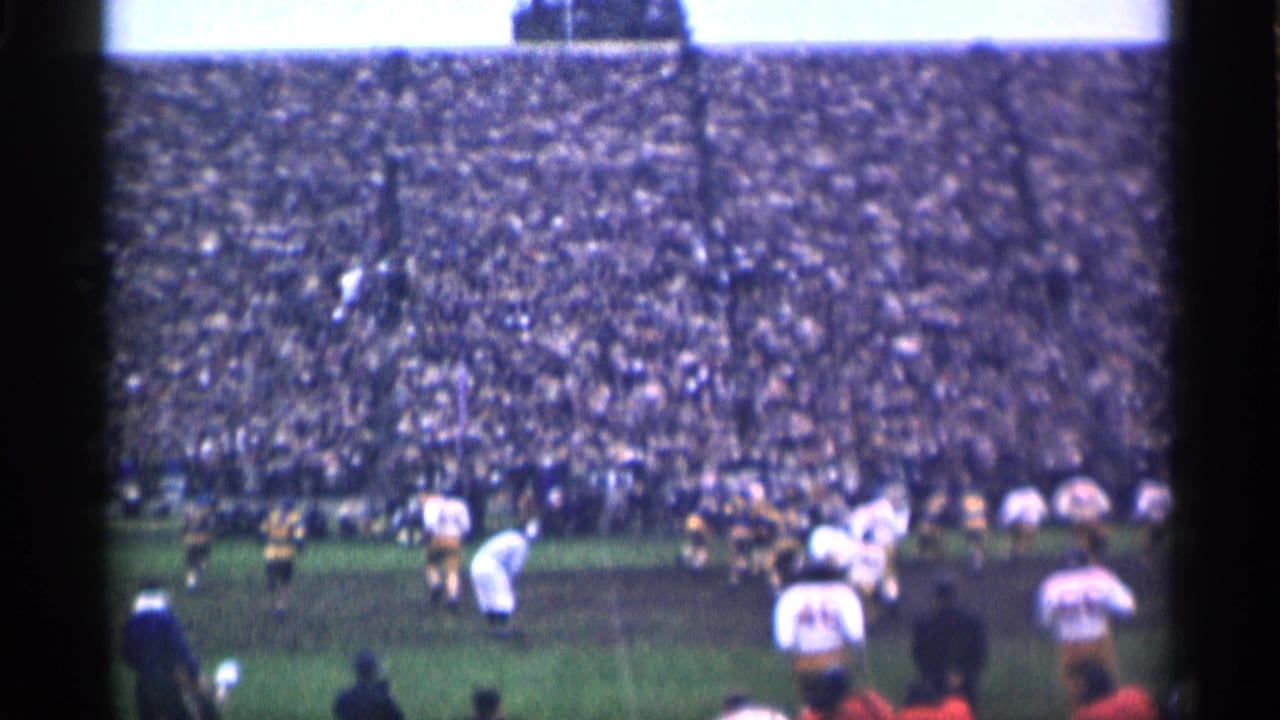 Wide stadium view of football players on a muddy field before a packed crowd.
