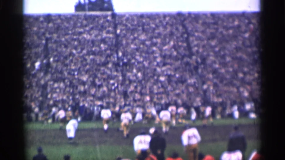 1940s home-movie frame of football players moving across a muddy San Francisco field with a dense stadium crowd behind them.