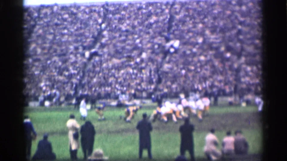 San Francisco 1944 football frame with sideline viewers in the foreground, players near the line, and crowded stands beyond.