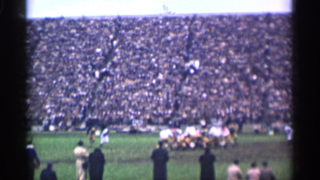 Football play on a green field with packed stadium stands behind it.