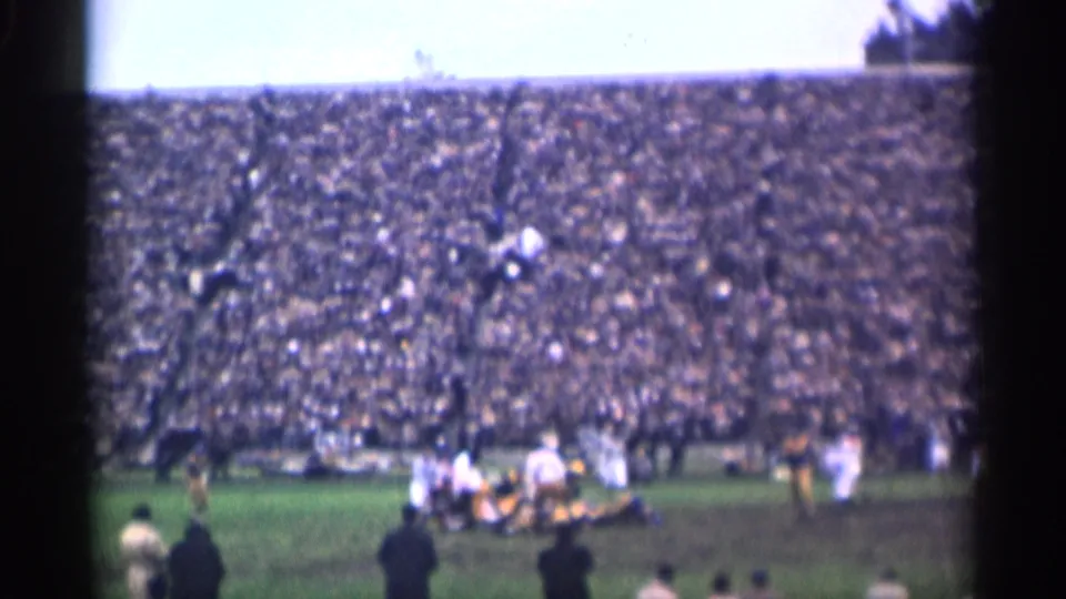 Distant 8mm view of a 1944 San Francisco football play with several players clustered on the field and spectators filling the stands.