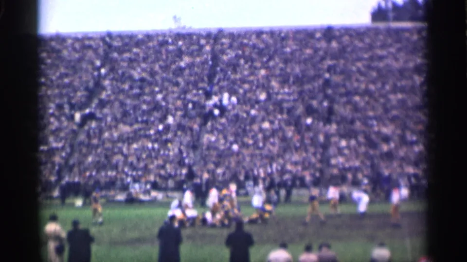 Home-movie frame of a 1944 San Francisco football game, with players gathered after contact and a dense crowd in the background.
