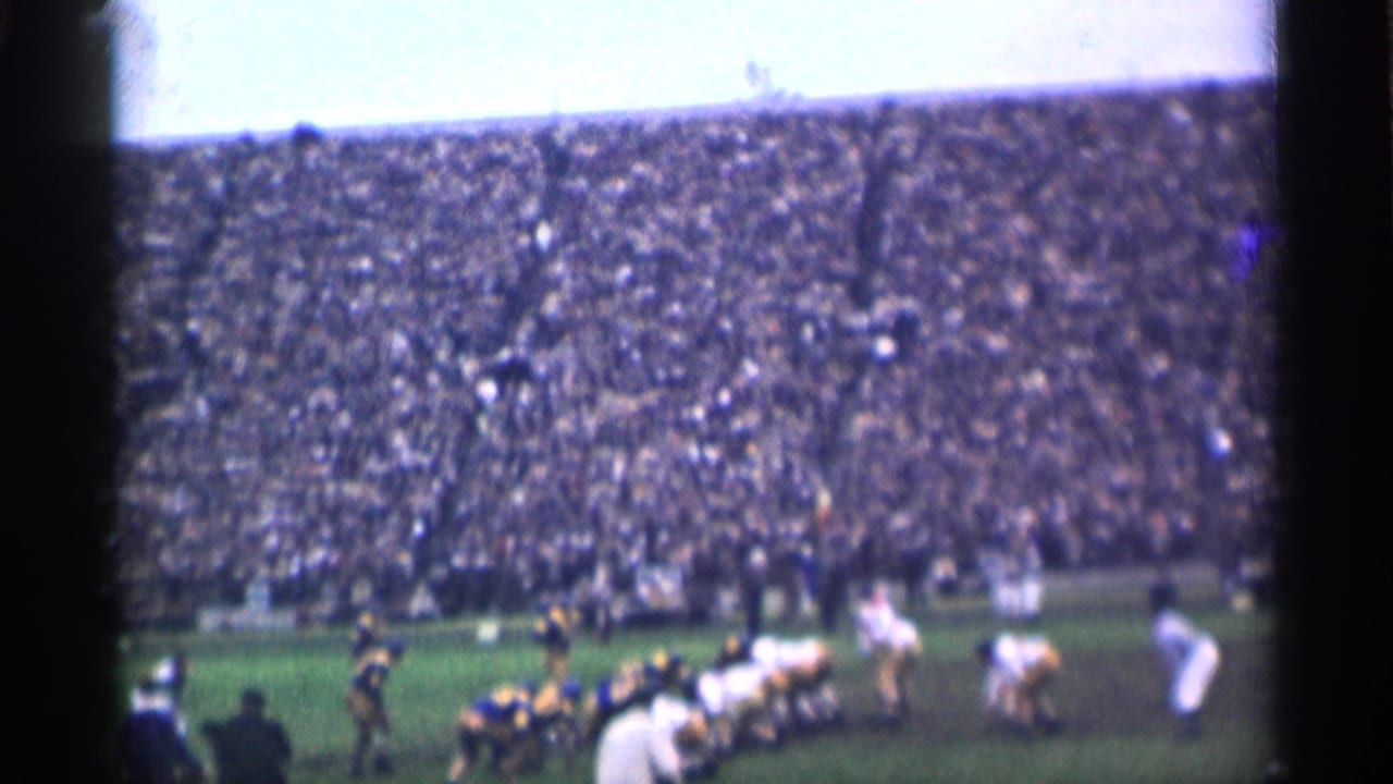 Football teams line up on a grass field before a play with a large crowd in the stands.