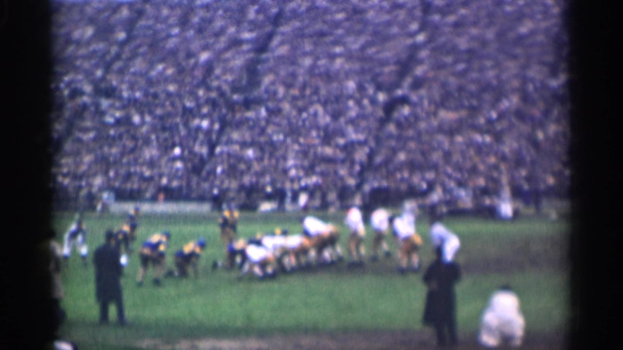 Two football teams face each other at the line with a dense crowd in the background.