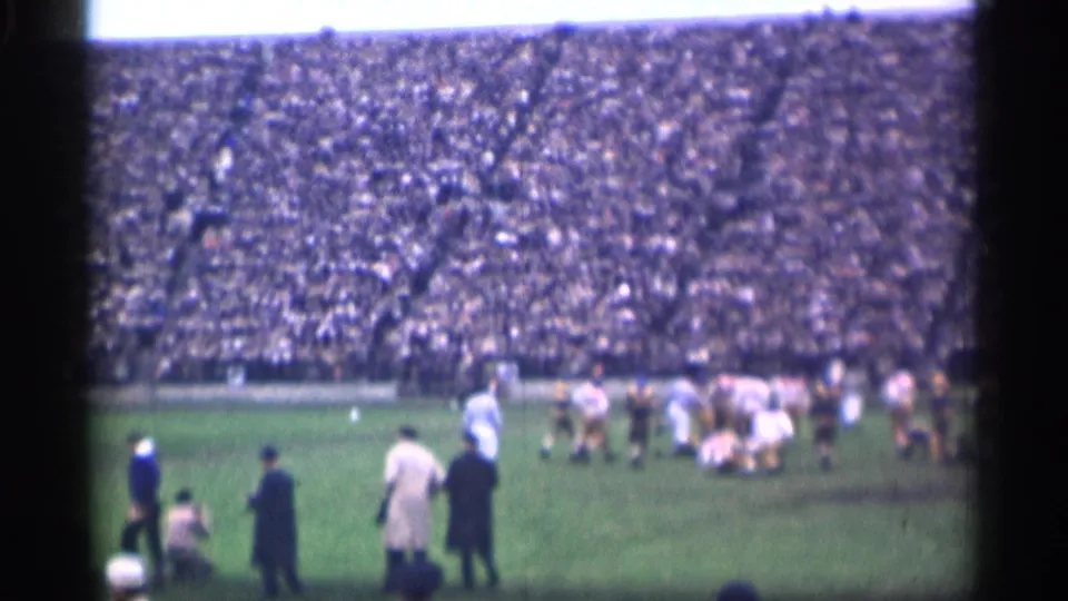 1944 San Francisco 8mm frame showing sideline spectators or staff in coats near a cluster of football players on a grass field.