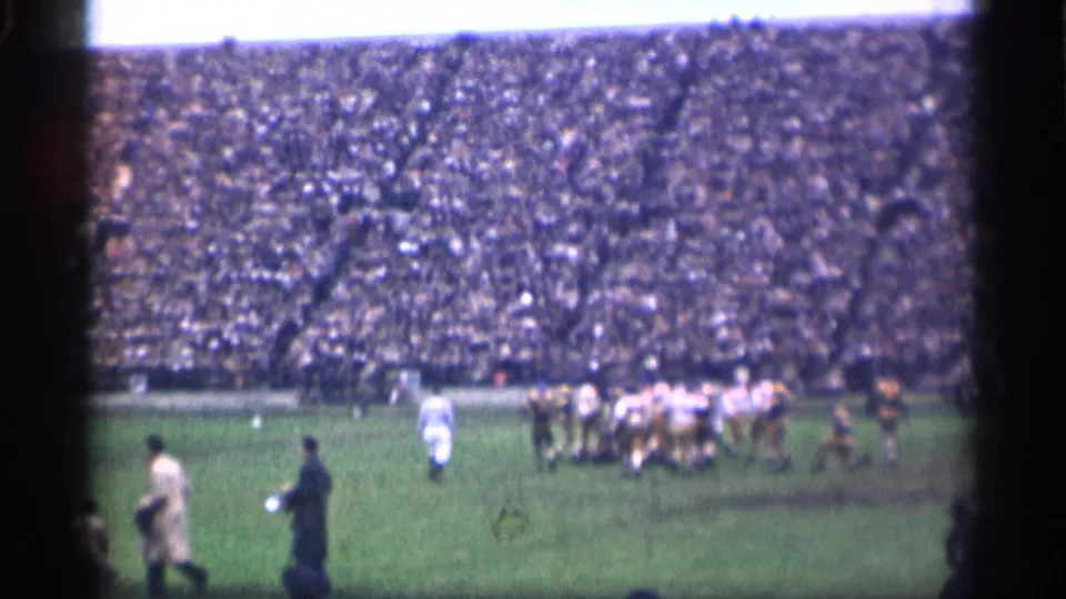 1944 San Francisco stadium frame with football players grouped on the field, sideline figures in coats, and packed spectator stands behind them.