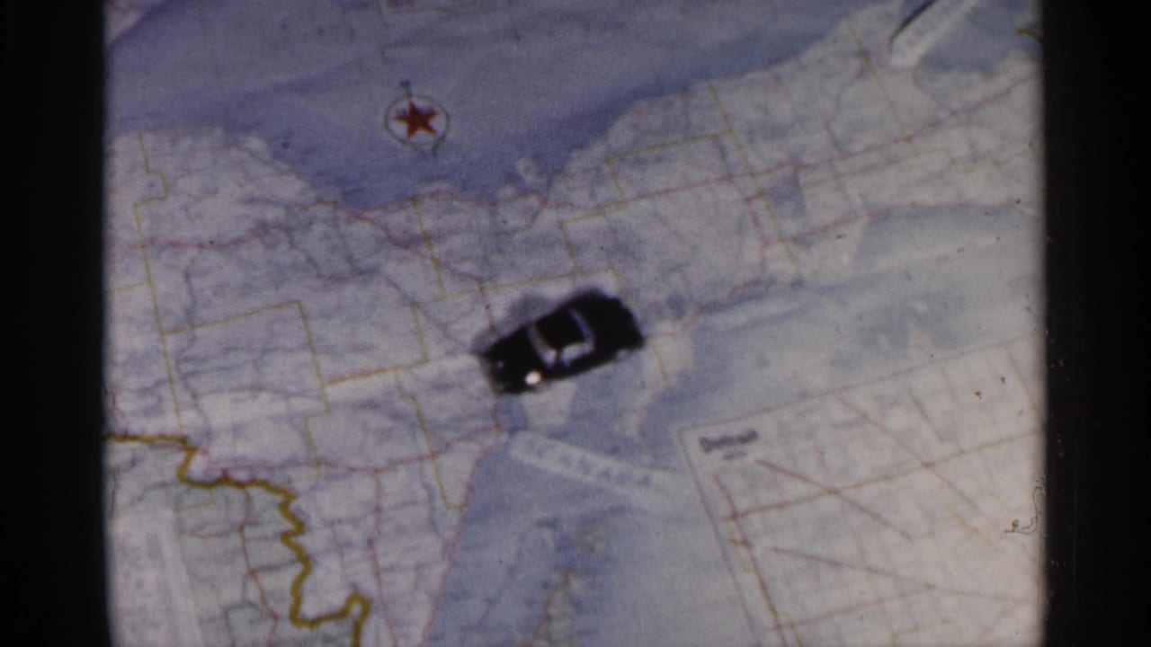 1960 8mm frame showing a small toy car placed on a paper map of Michigan near Great Lakes shoreline markings.
