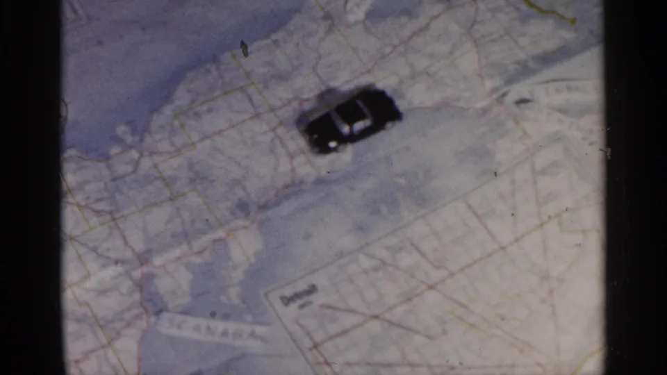 1960 8mm frame showing the small toy car on a paper map of Michigan, with road networks and water boundaries visible.