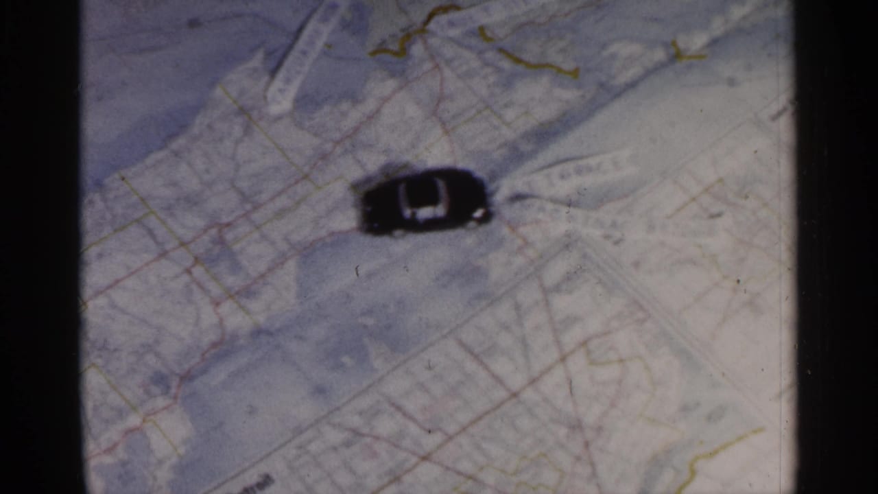 1960 Michigan-associated 8mm frame showing a small black toy car placed sideways on a printed paper map.