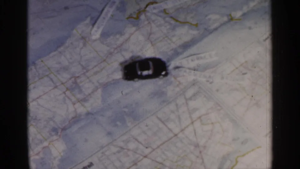 1960 Michigan-associated 8mm frame showing a small black toy car placed sideways on a printed paper map.