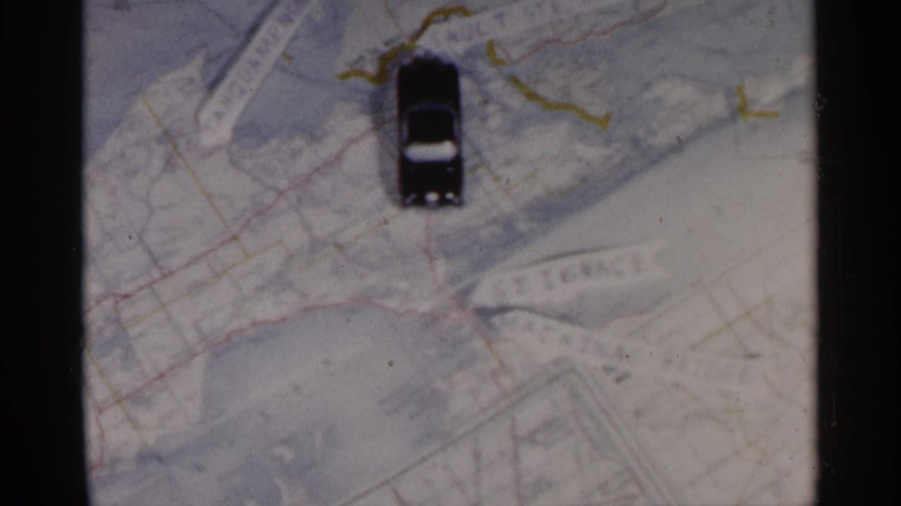 A black toy car sits on a paper map of Michigan, with drawn route lines and printed place names visible in 1960 8mm footage.
