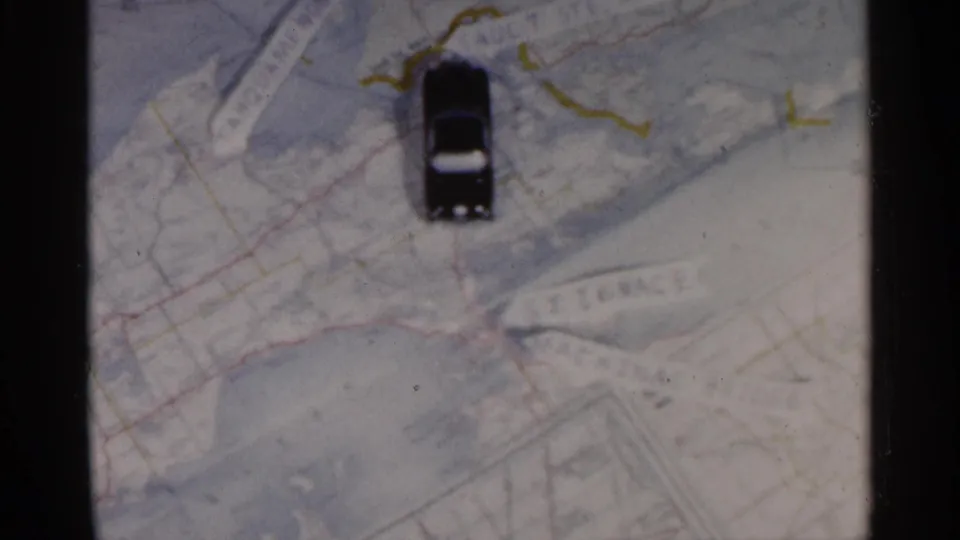 A black toy car sits on a paper map of Michigan, with drawn route lines and printed place names visible in 1960 8mm footage.