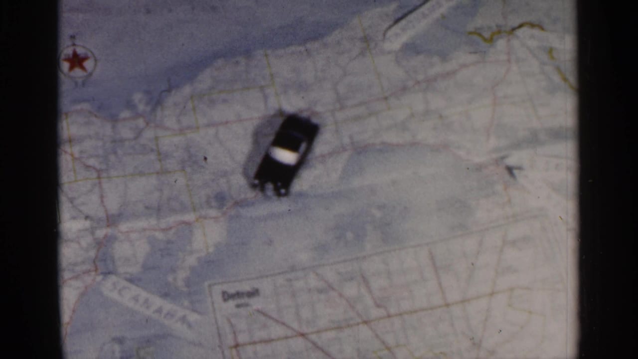 A small black car with a light roof sits on a printed Michigan road map, with Detroit visible in an inset area.