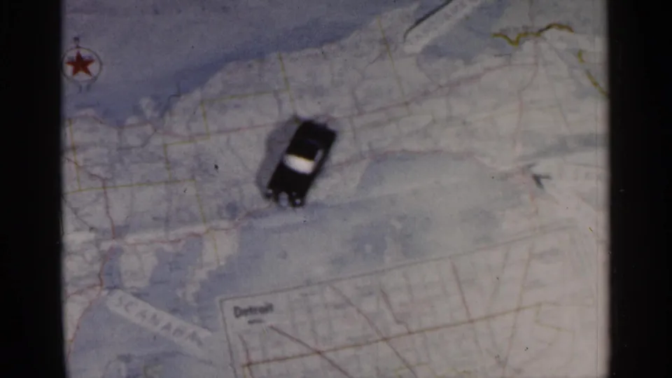 A small black car with a light roof sits on a printed Michigan road map, with Detroit visible in an inset area.