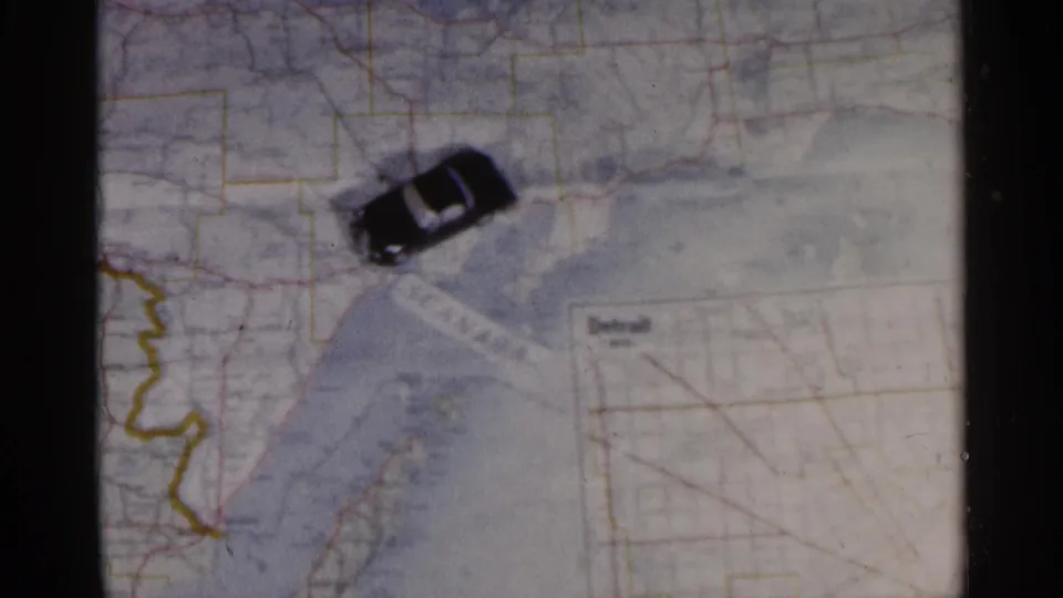 A black toy car sits over the lower portion of a Michigan road map, close to a Detroit inset and yellow boundary markings.
