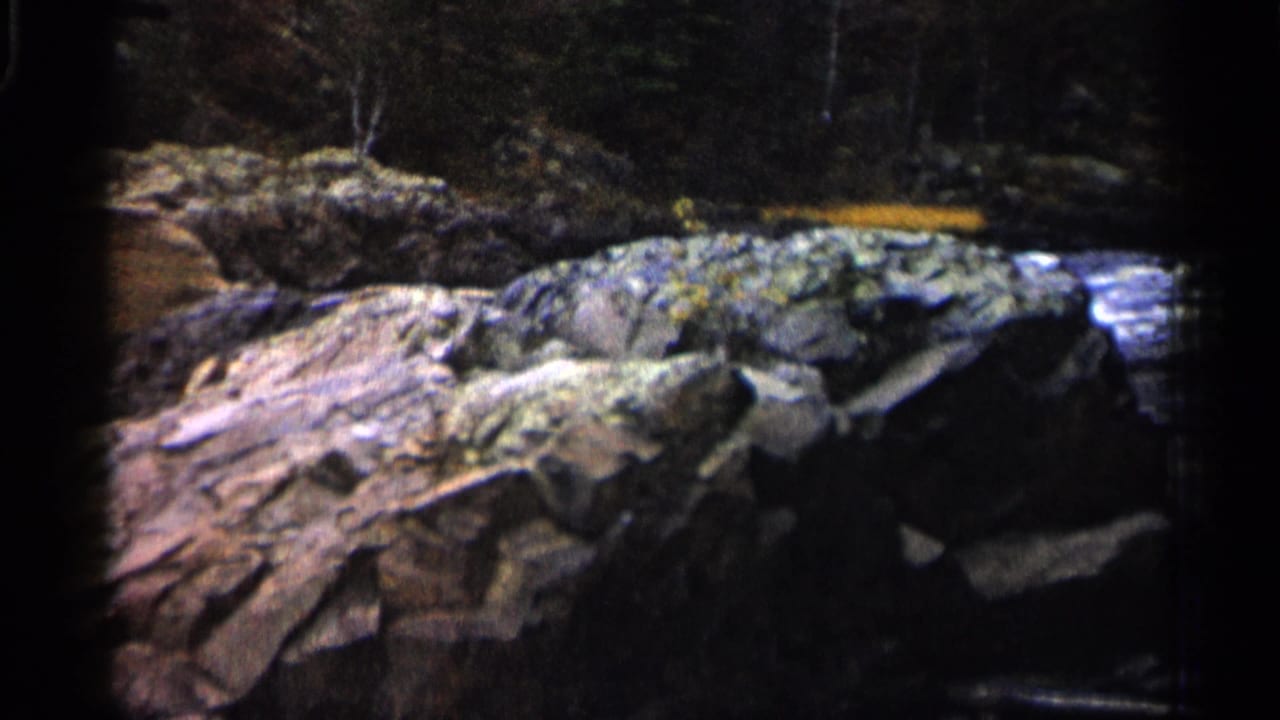 1960 Michigan 8mm frame showing layered rocks beside shallow river water with a dark wooded backdrop.