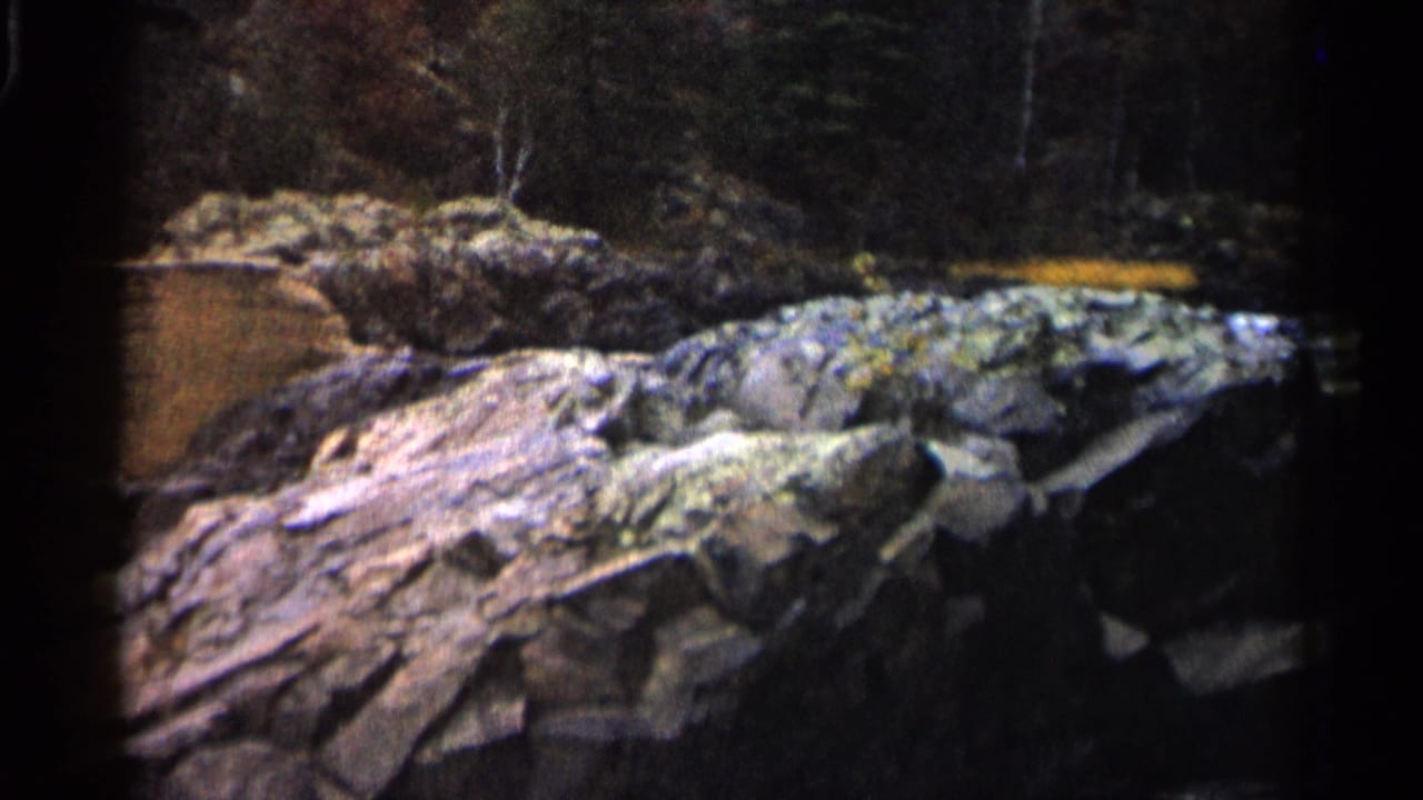1960 Michigan home-movie frame with a rocky ledge, scattered boulders, and water running behind the stones.
