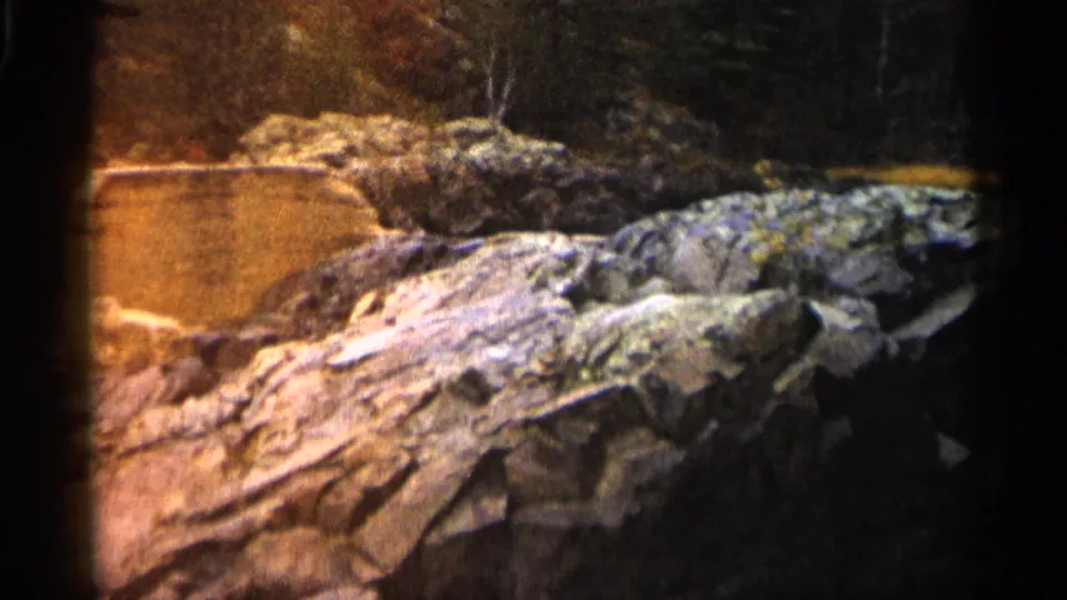 1960 Michigan 8mm frame showing warm exposed rocks beside shallow river water and a dark wooded background.