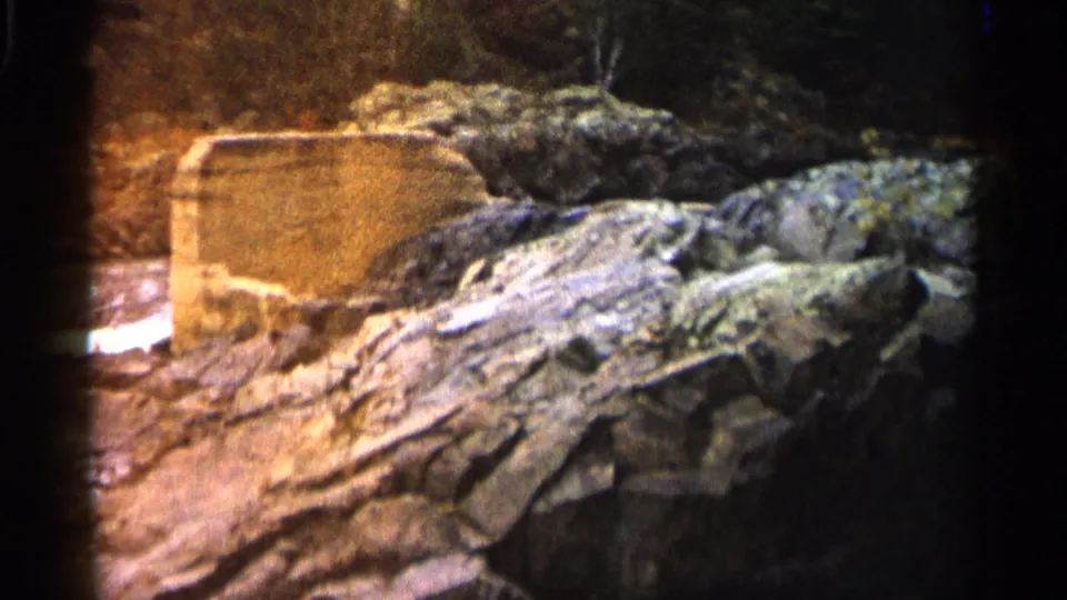 1960 Michigan home-movie frame showing a large flat rock formation next to flowing water with forest in shadow behind it.