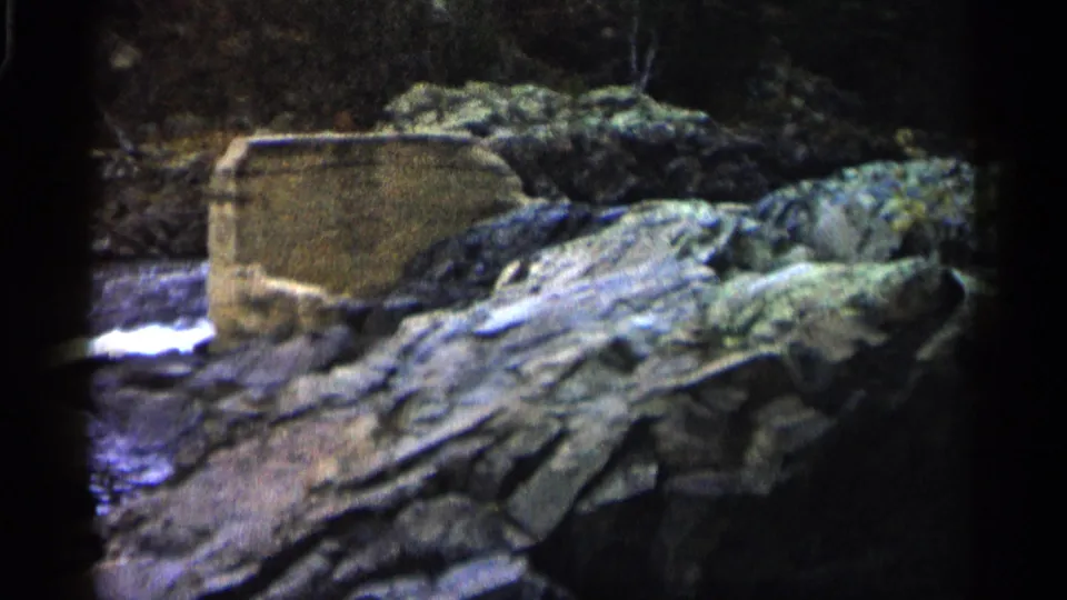 1960 Michigan 8mm frame showing a rocky riverbank, white water, and a worn concrete block beside layered stone.