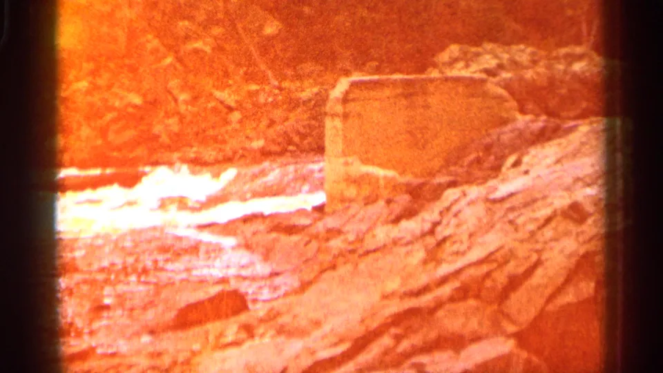 8mm frame with strong orange flare over a Michigan river scene, showing rocks, white water, and the same concrete structure.