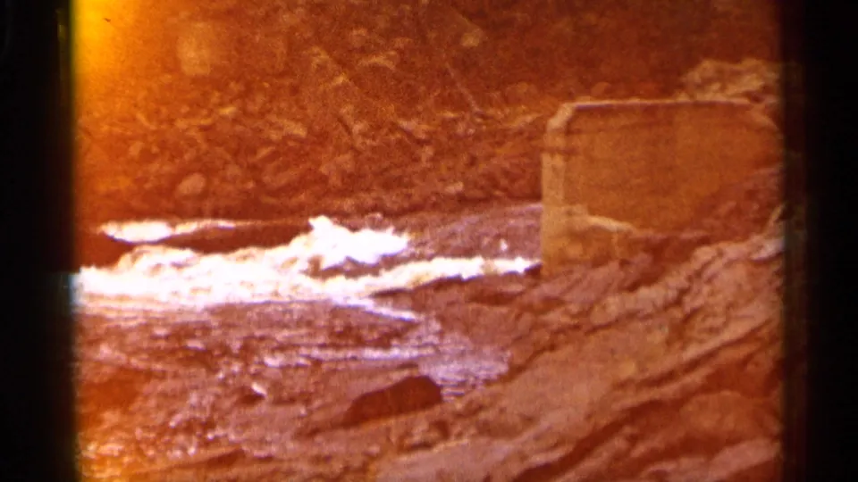 Final 1960 Michigan river frame with warm color shift, white water, layered rocks, and a concrete block partly at the right edge.