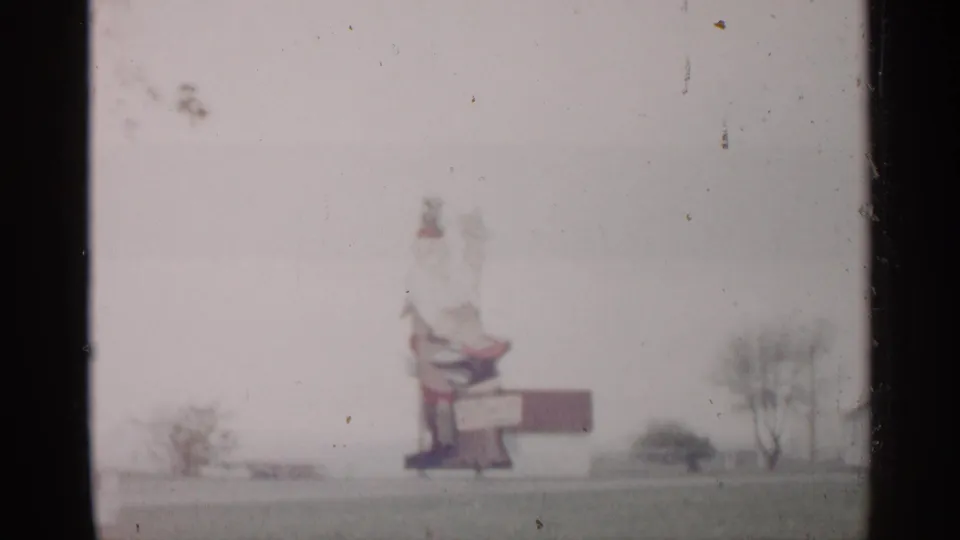 1960 Michigan 8mm frame showing a distant roadside sign-like structure with a bench-like shape, sparse trees, and washed-out sky.