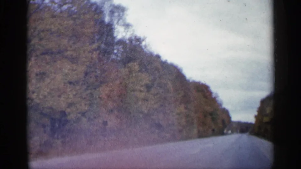 1960 Michigan 8mm frame from a moving vehicle, looking down a quiet road between wooded autumn embankments.