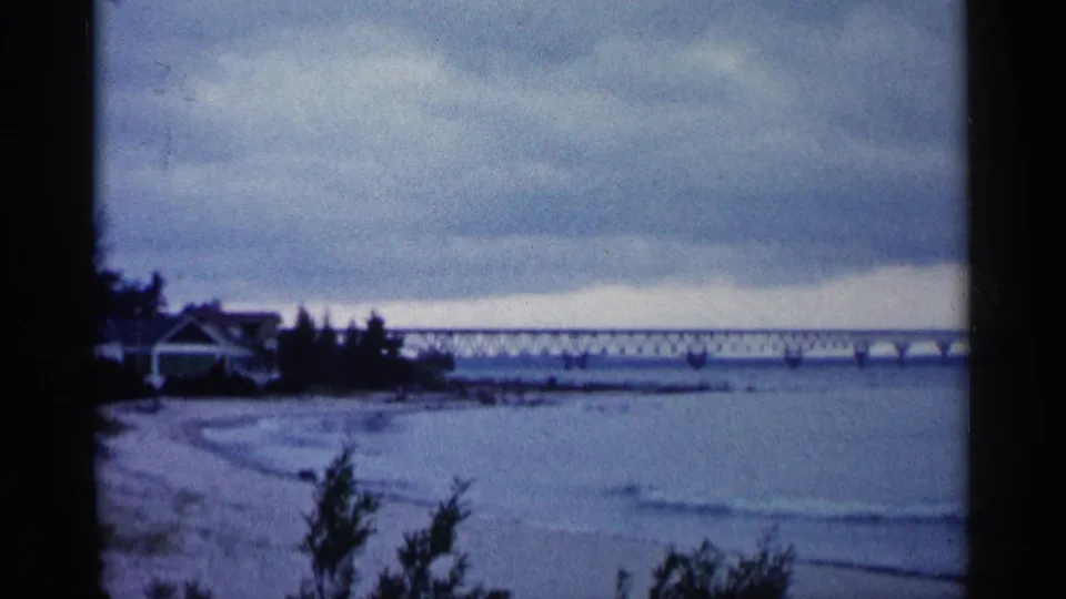 1960 Michigan home-movie frame showing lake water, a shoreline house, and a long bridge under a heavy overcast sky.