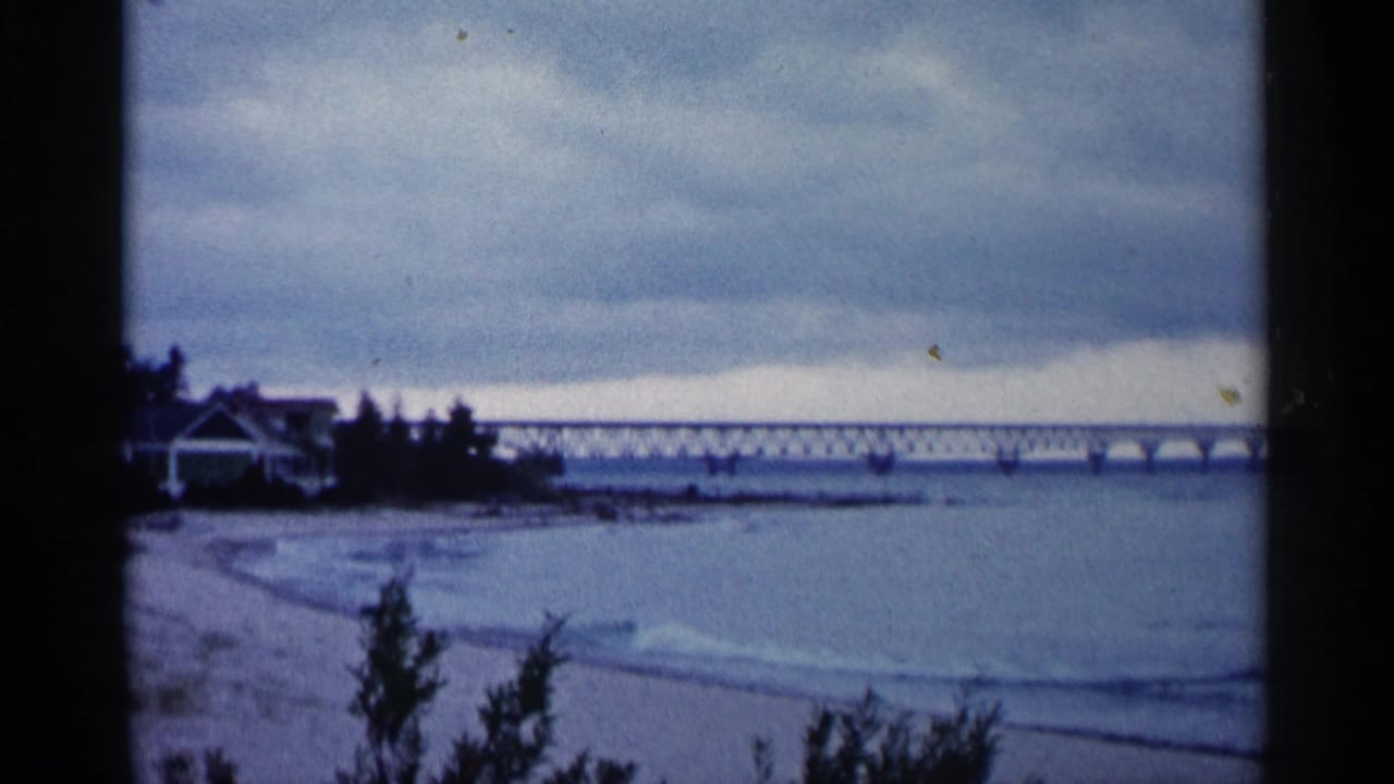 Overcast beach with gentle water, a long bridge in the distance, and trees at the left edge.
