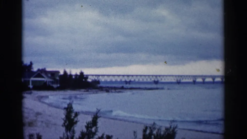 1960 Michigan 8mm view of a sandy shoreline, calm waves, dark foreground shrubs, and a long bridge stretching across the water.