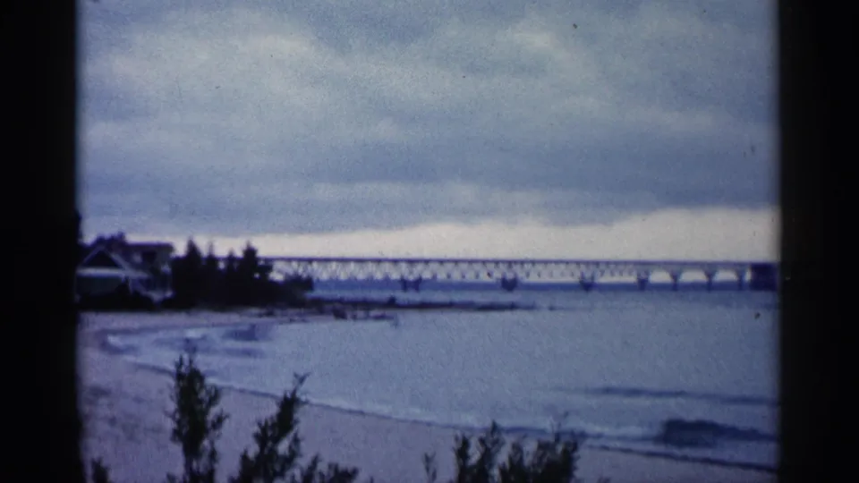 1960 Michigan shoreline frame showing a long bridge across calm water, muted waves, and trees beside a left-edge shoreline structure.